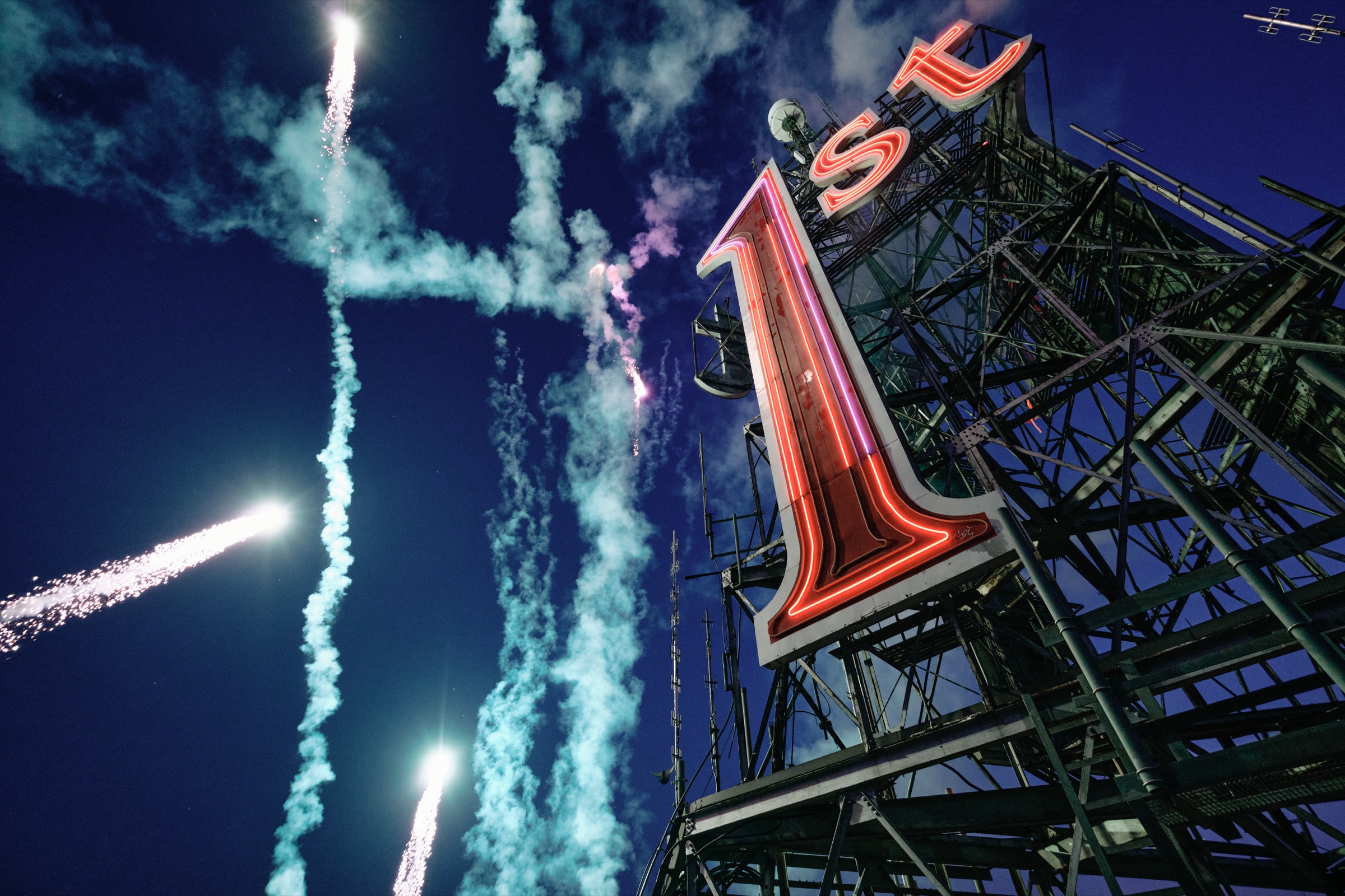 Fireworks streaking into the dusk sky beside the relit 1st sign